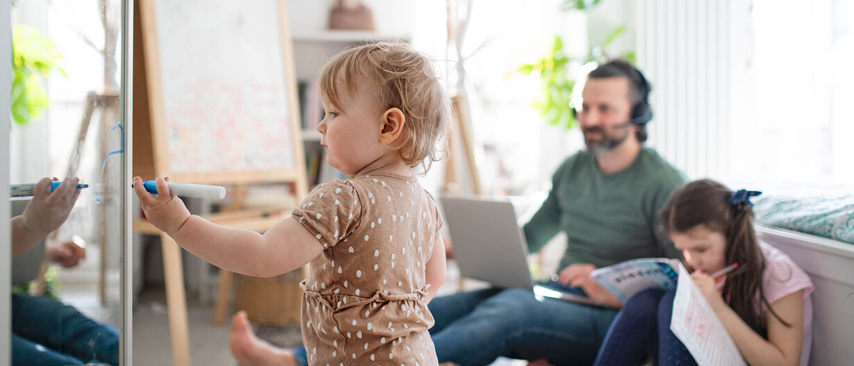 Kind spielt im Vordergrund, Vater arbeitet im Hintergrund während der Kinderbetreuung zu Hause